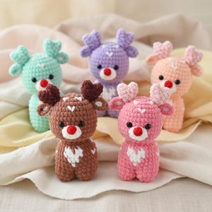 Könnte beinhalten: Fünf gehäkelte Rentierfiguren in Pastellfarben. Jedes Rentier hat herzförmige Geweihe, eine rote Nase und ein weißes Herzmuster auf der Brust. Die Farben sind Grün, Lila, Rosa, Pfirsich und Braun. Ideal als Dekoration.