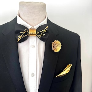 May include: A black suit with a white dress shirt, a black bow tie with gold branch accents, a gold lapel flower, and a gold pocket square. The bow tie features a gold band in the center.
