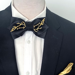 May include: A black bow tie with gold branch accents, paired with a matching gold pocket square. The bow tie is set against a white collared shirt and a black suit jacket, creating a formal and elegant look.