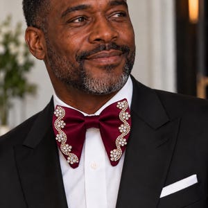 May include: A deep burgundy satin bow tie featuring intricate, wavy gold-toned embellishments adorned with clear sparkling crystals along both outer edges. The bow tie is worn over a crisp white button-down shirt and a classic black suit jacket.