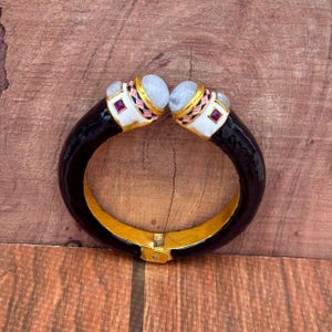 May include: A hinged cuff bracelet featuring a glossy black band with a gold-toned interior. The two ends are capped with white enamel, accented by small pink and blue geometric patterns, square red stones, and rounded, translucent grey cabochons.