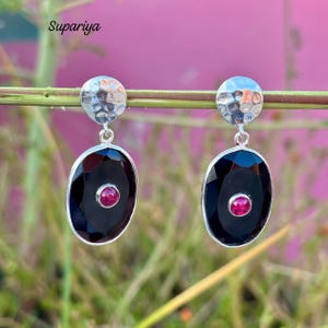 Peut inclure: Boucles d'oreilles en argent avec une texture martelée en haut. Chaque boucle d'oreille présente une grande pierre ovale noire facettée avec une petite pierre rouge ronde au centre. La marque "Supariya" est visible en haut à gauche.