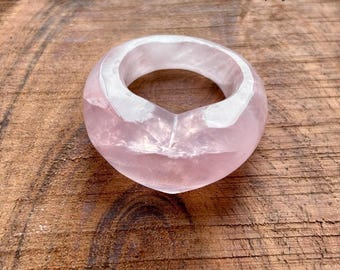 Pode incluir: Um anel grosso em forma de coração feito de quartzo rosa translúcido. O anel tem um acabamento liso e polido e uma faixa de quartzo branco. O anel é exibido em uma superfície de madeira.