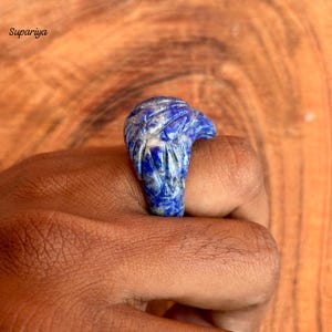 May include: A carved blue lapis lazuli ring in the shape of a bird's head. The ring is held up by a hand. The ring has white veining throughout the blue stone. The word "Supariya" is in the top left corner.