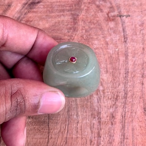 May include: A light green jade ring with a rounded top and a small, round, red gemstone set in a gold-colored bezel. The ring is held against a wooden surface.