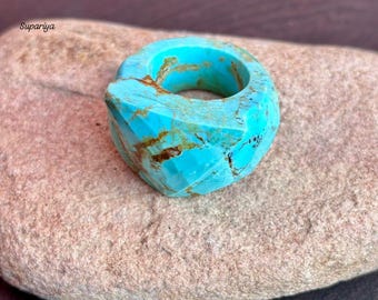 May include: A faceted turquoise ring with a geometric design. The ring is a vibrant blue with brown veining, set against a neutral stone surface. The ring's unique cut and color make it a striking piece of jewelry.