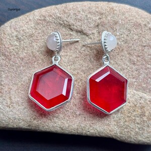 May include: A pair of silver earrings. Each earring has a small, round, pale pink stone at the top, a silver setting, and a large, faceted, hexagonal, red gemstone pendant. The earrings are displayed on a textured, tan surface.