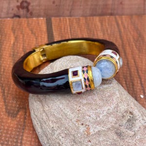 May include: A hinged bangle bracelet featuring a dark brown, glossy, curved band with a gold-tone interior. The ends are decorated with white enamel, small square red and blue stones, and large, round, translucent, pale blue cabochon gemstones.