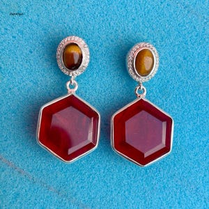 Puede incluir: Un par de pendientes plateados con diseño de piedras preciosas. Cada pendiente presenta una piedra ovalada marrón en la parte superior, rodeada de pequeñas piedras transparentes. Una gran piedra roja en forma de hexágono cuelga debajo.