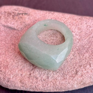 May include: A faceted, light green gemstone ring with a square face. The ring is set on a pink, textured surface. The word "Supariya" is in the upper left corner.
