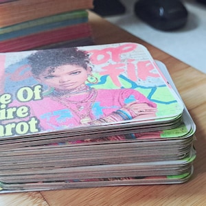 Op de afbeelding: Een stapel tarotkaarten met een kleurrijk ontwerp en de tekst "Ace Of Fire Tarot". De kaarten tonen een portret van een persoon die een roze shirt draagt. Andere stapels kaarten zijn zichtbaar op de achtergrond.