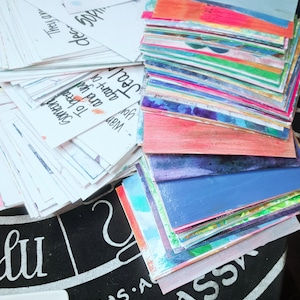 Pode incluir: Uma coleção de cartões de arte coloridos pintados à mão e notas manuscritas. Os cartões apresentam vários designs e cores abstratas, incluindo azul, laranja e rosa. Algumas notas têm o texto "They're a Kitchen Witch Spells bound to backfire."