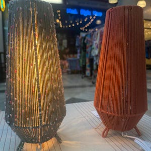 May include: Two decorative lamps with a conical shape. One lamp is gray with pink accents, and the other is orange. Both lamps are made of yarn and have three small legs. The lamps are lit, and the background is blurred.
