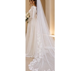 Peut inclure: Un long voile de mariée blanc et transparent orné de broderies florales complexes en dentelle sur les bords et d'un ourlet festonné. Le voile est composé d'un tissu en tulle léger qui tombe jusqu'au sol, révélant des motifs floraux détaillés.