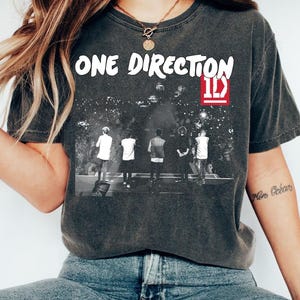 May include: Dark grey t-shirt with the white text "ONE DIRECTION" above a black and white concert image. The shirt features the "1D" logo in a red box. The t-shirt is made of a soft material.