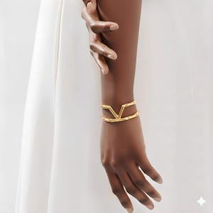 May include: A gold-coloured bracelet featuring a V-shaped design. The bracelet is displayed on a wrist, with the arm and hand in view. The background is a soft white, accentuating the jewellery's design and colour.