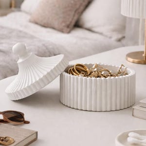 Può includere: Un portagioie in ceramica bianca con base cilindrica scanalata e coperchio rimovibile abbinato con pomello sferico. Il contenitore è riempito con varie catenine, anelli e braccialetti in metallo dorato, caratterizzato da un esterno testurizzato.