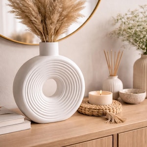 Pode incluir: Um vaso de cerâmica branco com um design circular e um buraco no centro, preenchido com capim-dos-pampas seco. Uma vela acesa, uma tigela pequena e outros vasos decorativos estão em uma superfície de madeira. Um espelho com moldura dourada está no fundo.