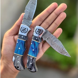 May include: Two folding knives with patterned steel blades and blue and gray handles. The handles have a blue, gold, and white design. Each knife has a silver bolster with a logo. One knife is open, and the other is closed.