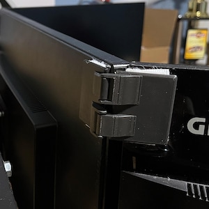 以下が含まれることがあります： 黒いモニターマウント。2つの調整可能なクリップが付いており、黒いコンピューターモニターの背面に固定されています。モニターの前面には「GIGABYTE」のブランド名が印刷されています。マウントは3Dプリントされているようです。