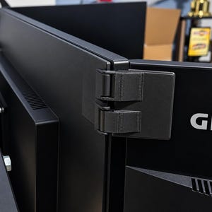 Puede incluir: Un conector de bisagra para monitor de plástico negro que une dos pantallas de ordenador. El soporte presenta una superficie texturizada y un diseño de doble bisagra, fijando de forma segura los paneles traseros de los monitores para una configuración.