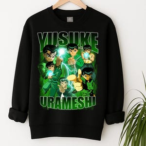Peut inclure: Sweat-shirt noir avec un motif du personnage de l'anime Yu Yu Hakusho, Yusuke Urameshi. Le design vert affiche le nom du personnage et plusieurs images en action. Le sweat-shirt est suspendu à un cintre en bois.