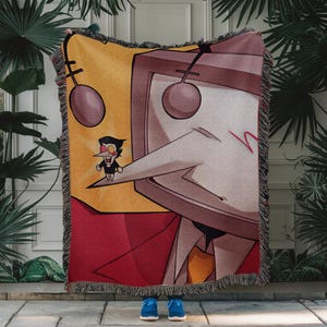 May include: A woven throw blanket featuring a cartoon character with a large screen head, a long nose, and a small figure standing on the nose. The blanket has a yellow, red, and beige color scheme with a fringed edge.