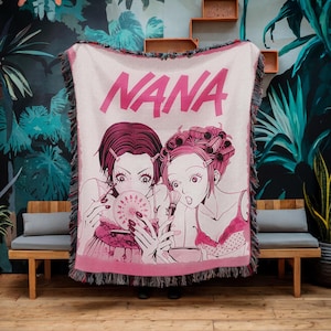 May include: A pink and white woven throw blanket featuring the word "NANA" in large pink letters. The blanket displays an anime-style illustration of two women. The blanket has a fringed edge and is displayed against a backdrop of tropical foliage.