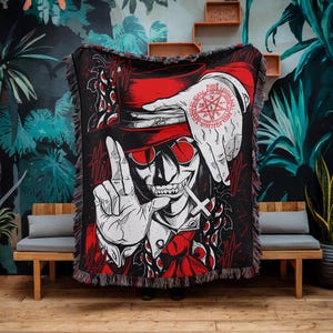 May include: Black woven throw blanket featuring a red-hatted, sunglasses-wearing skull graphic with a cross and a hand with a pentagram tattoo. The design is in red and white on a black background, with fringed edges.