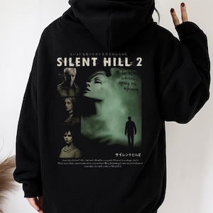 Puede incluir: Sudadera con capucha negra con un gráfico de Silent Hill 2. El diseño incluye imágenes de personajes y el texto "SILENT HILL 2" en blanco. La parte posterior presenta un diseño verde y blanco con las palabras "The door that opens into darkness, opening into nightmares."
