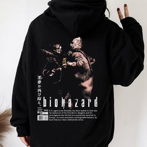 Może przedstawiać: Czarna bluza z kapturem z grafiką "Biohazard". Projekt zawiera postarzone zdjęcie dwóch postaci, japoński tekst i tytuł gry. Bluza ma kaptur ze sznurkiem i kieszeń typu kangur.