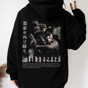 Może przedstawiać: Czarna bluza z kapturem z grafiką z gry wideo Biohazard. Projekt zawiera japoński tekst i obrazy postaci. Bluza ma kaptur ze sznurkiem i długie rękawy.