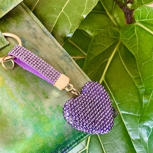 May include: A keychain featuring a purple heart-shaped charm covered in small sparkling crystals. The charm is attached to a matching purple strap encrusted with crystals and a gold-toned metal ring, creating a cohesive and decorative accessory.