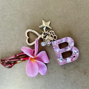 May include: A gold-toned keychain with a star and heart-shaped clasp, a pink floral accent, and a pink letter "B" charm encrusted with rhinestones. The keychain is on a neutral background.