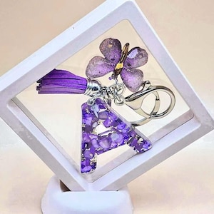 May include: A purple keychain featuring the letter 'A' filled with purple stones, a purple tassel, and a butterfly charm. The keychain is attached to a silver clasp. The items are displayed in a white, angled box.