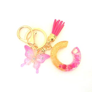 Könnte beinhalten: Ein goldfarbener Schlüsselanhänger mit einem rosafarbenen, durchscheinenden Schmetterlingsanhänger mit kleinen klaren Strasssteinen, einem Buchstaben-C-Anhänger mit Goldglitter und rosa Harzfragmenten sowie einer leuchtend rosa Quaste.