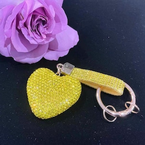 Pode incluir: Um chaveiro amarelo em forma de coração coberto por pequenos cristais brilhantes. O chaveiro tem uma alça amarela combinando e um porta-chaves prateado. Uma rosa roxa está no fundo sobre uma superfície escura.
