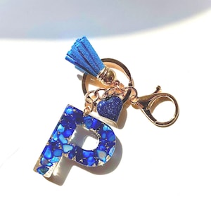Puede incluir: Un llavero dorado con una borla azul y un dije con la letra "P". La "P" está rellena de piedras azules y doradas. Un pequeño dije de corazón azul cuelga debajo de la borla. El llavero tiene un cierre.