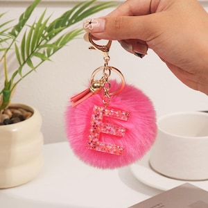 May include: A pink fluffy keychain with a gold-colored metal clasp and a tassel. The keychain features a large, glitter-filled letter "E" in the center. The keychain is held by a person's hand.