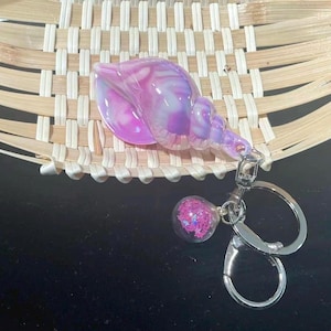 May include: A purple and white seashell-shaped keyring with a silver clasp and a small, round charm filled with pink glitter. The keyring is set against a woven, light brown background.