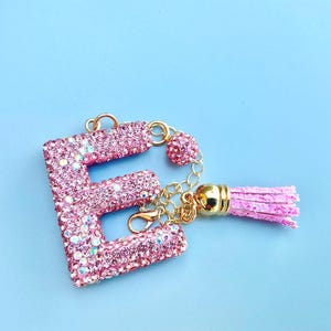Könnte beinhalten: Rosa Buchstabe "E" Schlüsselanhänger, besetzt mit schillernden Strasssteinen. Die goldfarbene Hardware umfasst einen Karabinerhaken, einen herzförmigen Anhänger und eine Quaste. Die Quaste ist hellrosa.