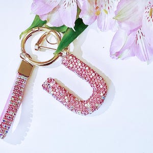 May include: A pink keychain with a gold ring and a matching letter "J" charm, both encrusted with sparkling pink rhinestones. The keychain has a pink leather strap. Soft pink flowers are in the background.