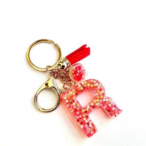 May include: A pink letter "R" keychain with gold star-shaped glitter. The keychain has a gold clasp, a red tassel, and a small clear bead with red glitter. The keychain is on a white background.