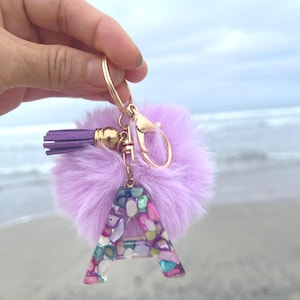 Op de afbeelding: Een decoratieve sleutelhanger met de letter "A" in een kleurrijk mozaïekontwerp. De sleutelhanger heeft een pluizige, lichtpaarse pompom, een kleine paarse kwast en goudkleurige hardware. Een gepersonaliseerd accessoire.