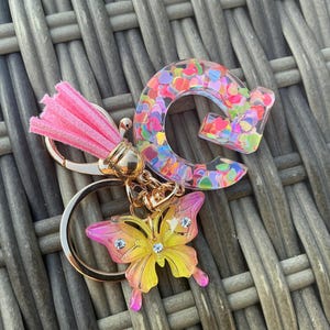 Op de afbeelding: Een sleutelhanger met een goudkleurige metalen ring, een roze kunstsuède kwastje, een doorzichtige letter G van hars gevuld met veelkleurige hartvormige confetti en een geel-roze vlinderbedel versierd met drie kleine doorzichtige strass-steentjes.