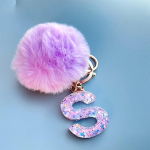 May include: A fluffy, lavender and pink pom-pom keyring with a gold-coloured metal ring. The keyring features a glittery, light purple letter "S" charm with iridescent sparkles. The background is a solid light blue.