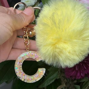 Puede incluir: Un llavero dorado con un pompón amarillo esponjoso y un dije con la letra "C" lleno de brillantina. El dije tiene una base de resina transparente con brillantina colorida. Una pequeña campana dorada cuelga de la cadena.