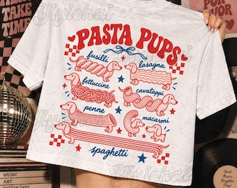 Puede incluir: Una camiseta blanca con un estampado gráfico en rojo y azul titulado PASTA PUPS. El diseño muestra varios perros salchicha con formas de tipos de pasta, incluyendo fusilli, lasagna, fettuccine, cavatappi, penne, macaroni y spaghetti.