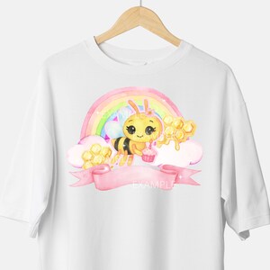 Op de afbeelding: Wit T-shirt met een cartoon bij die een cupcake vasthoudt, tegen een regenboog- en honingraatontwerp. Een roze banner met het woord "EXAMPLE" staat eronder. Het T-shirt hangt aan een houten hanger.