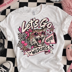 Puede incluir: Una camiseta blanca de manga corta con un gráfico de un gato montés con gafas de sol en forma de estrella. El diseño incluye el texto "Let's Go" y "WILDCATS" en un megáfono, con lazos rosas, cerezas, estrellas y un patrón de cuadros.